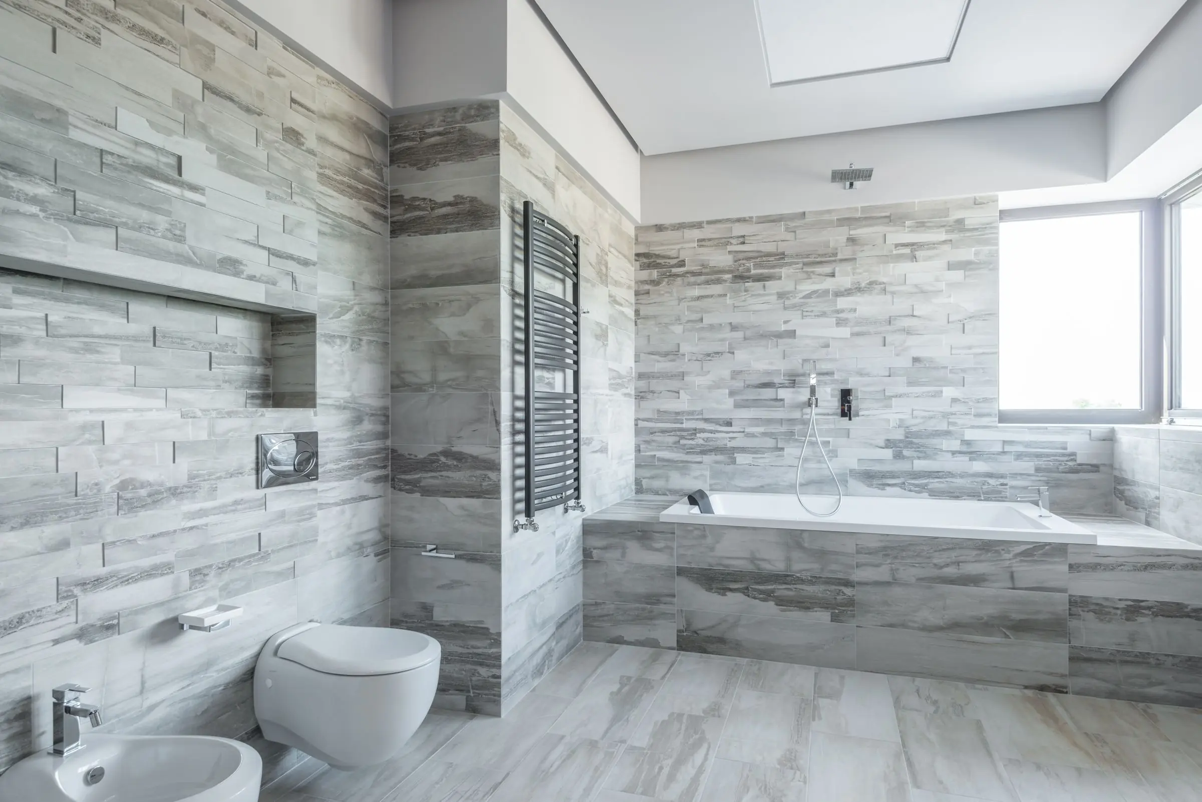 Une salle de bains carrelée de gris du sol au plafond, avec une baignoire et une grande fenêtre
